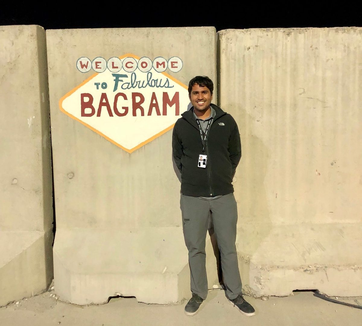 Vinoo Ganesh 站在阿富汗 Bagram 空军基地一面画着「Welcome to Fabulous Bagram」的混凝土墙前